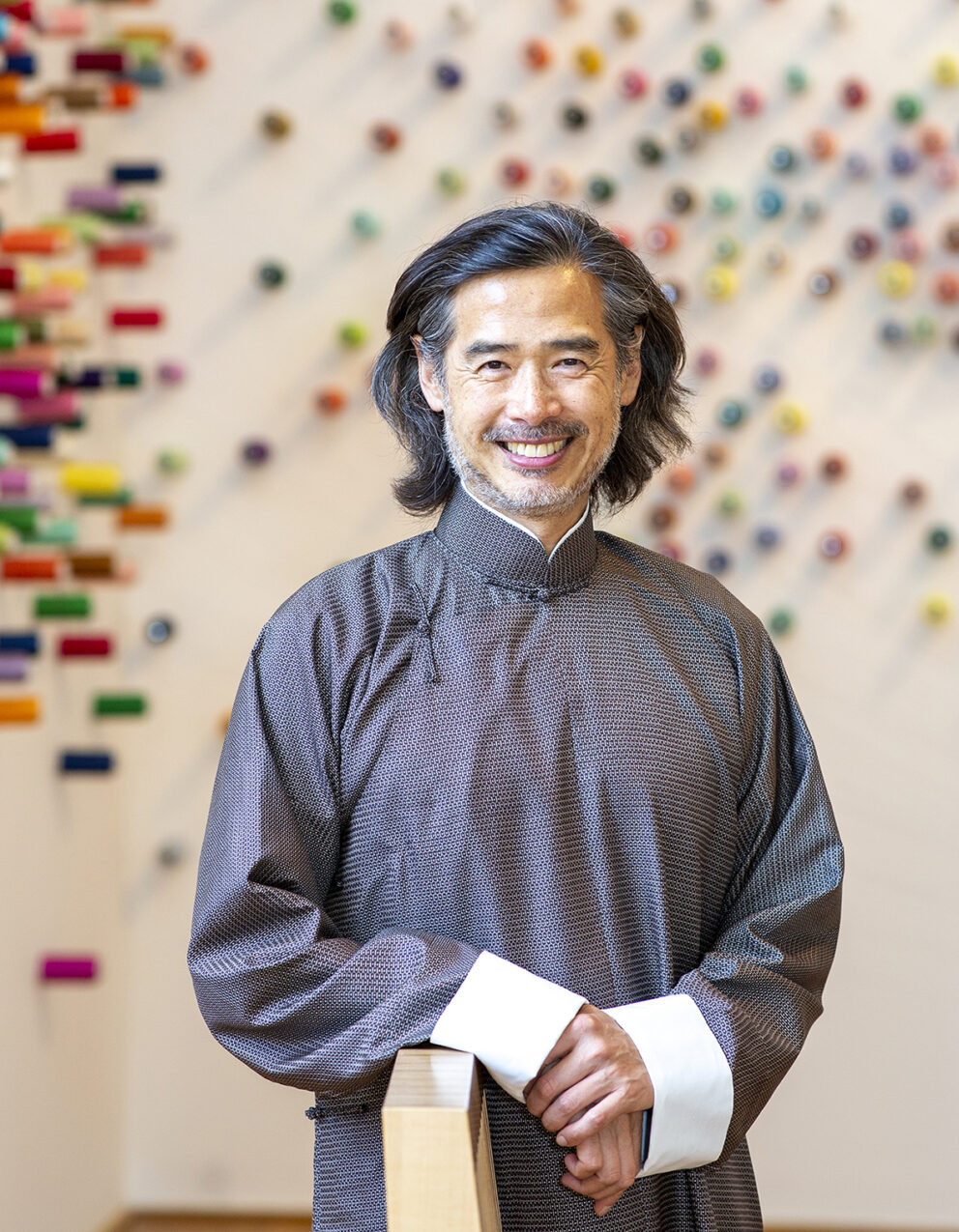 Photograph of Lee Mingwei, from the waist up. One of his artworks is pictured blurred in the background.